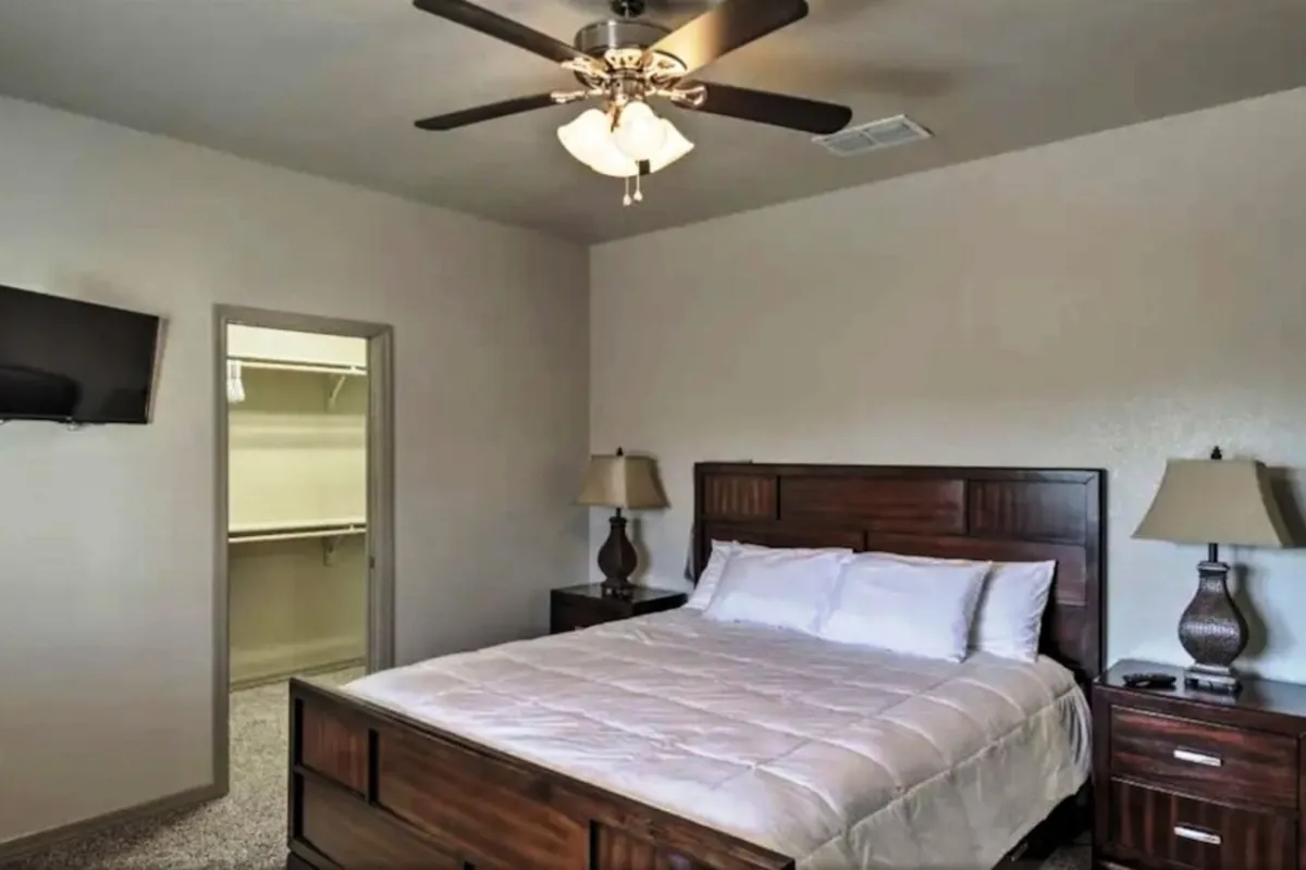 A tranquil bedroom featuring a bed with crisp white linens, wooden nightstands on either side, a ceiling fan with a light fixture above, a television mounted on the wall, and a closet door. Soft morning light fills the room, creating a welcoming space for restful sleep or quiet reading.