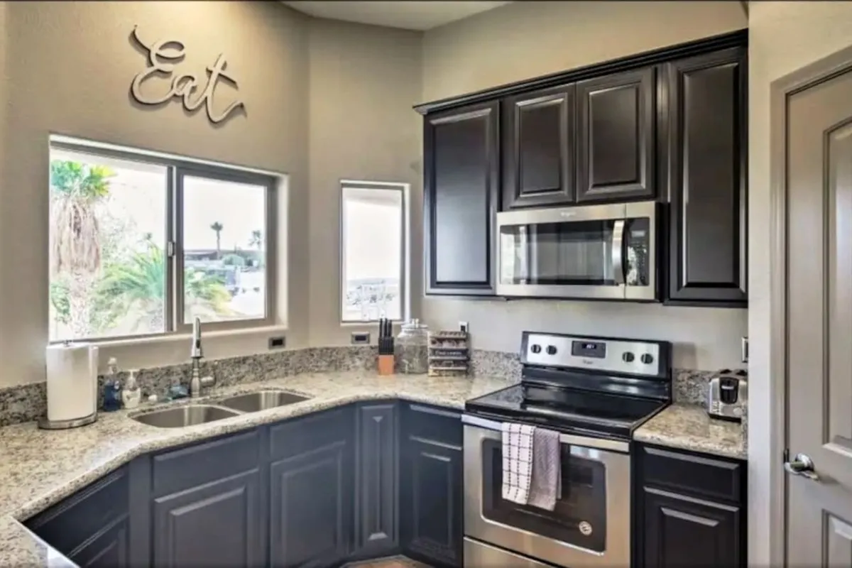 This image captures a view of the kitchen, showcasing granite countertops, stainless steel appliances, and a welcoming 'Eat' sign near a window that offers a pleasant outside view. The scene conveys the kitchen's role as a vibrant gathering spot for cooking and sharing meals.