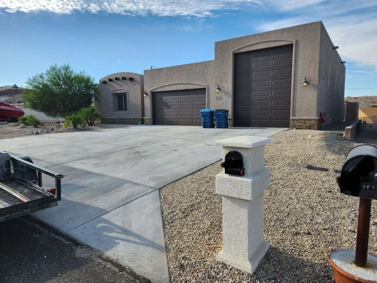 A sunlit exterior shot of a single-family home in a quiet Lake Havasu City neighborhood. The image captures the welcoming facade, dual garage doors, neat driveway, and a classic mailbox, all framed by desert landscaping. The setting suggests ample parking and a sense of privacy and space.