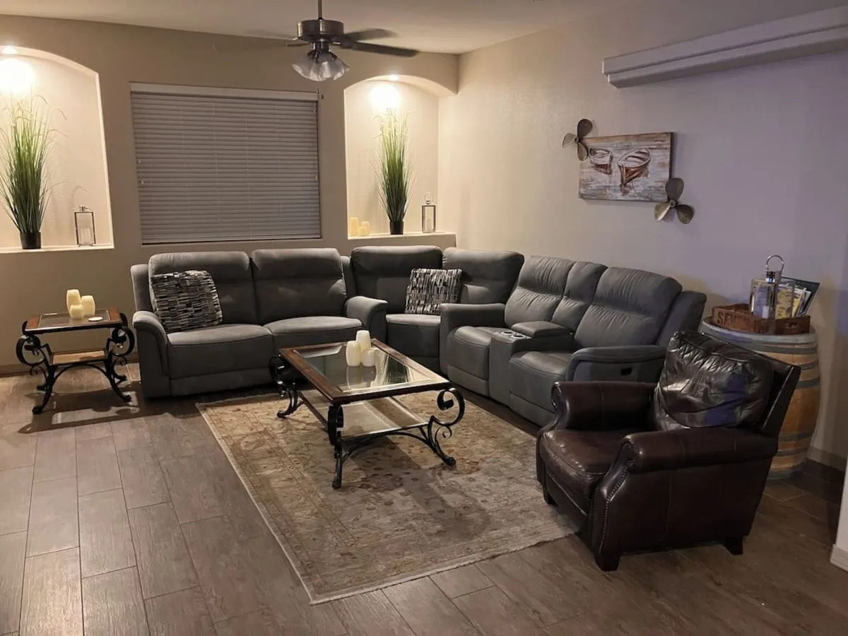 A cozy interior living area features a grey sectional sofa, glass coffee table, decorative wall art, and a barrel side table. The space is designed for socializing, relaxing, and sharing laughter after a day outdoors.