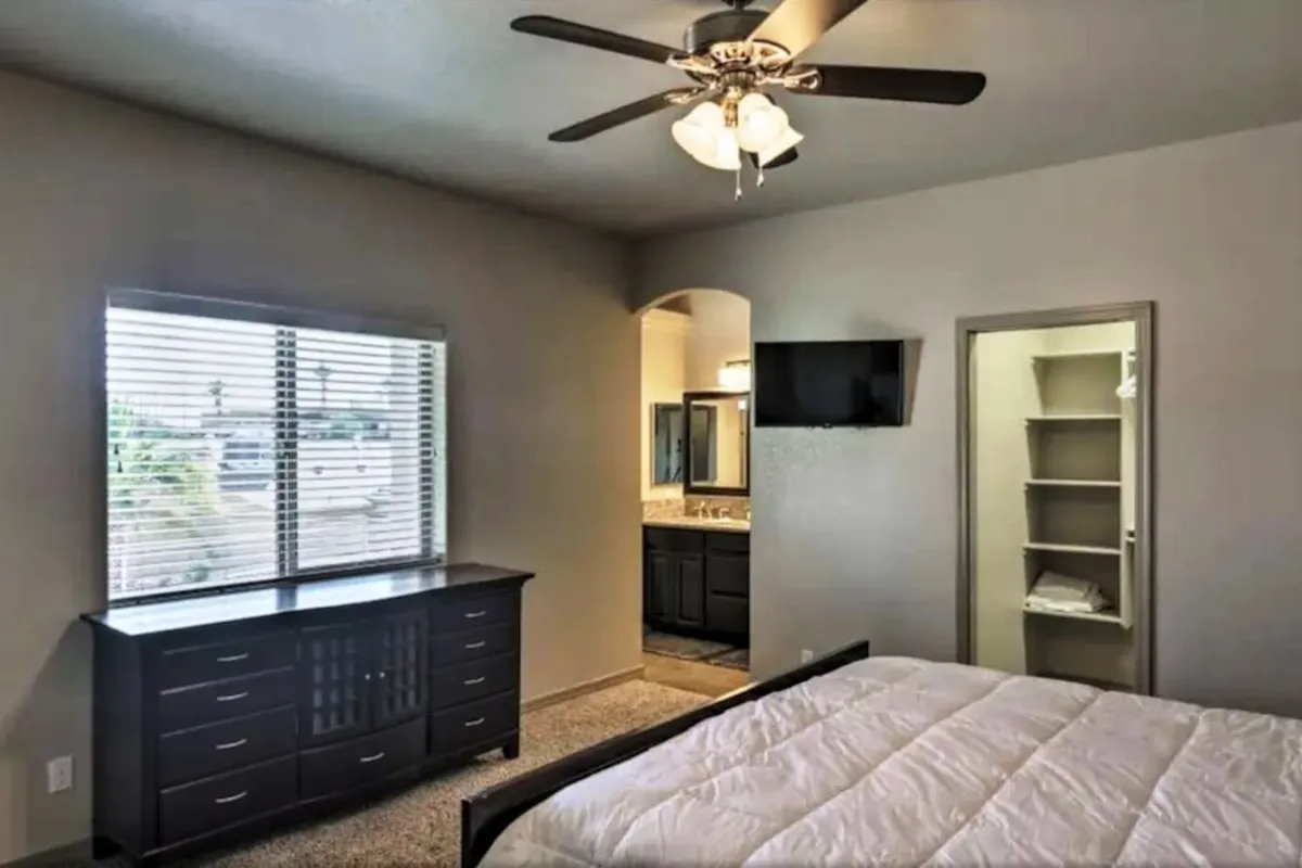 A cozy bedroom with crisp white bedding, a dark wood dresser, and a vanity area with a mirror. The room is bathed in gentle natural light from a window, creating a comfortable and restful atmosphere.