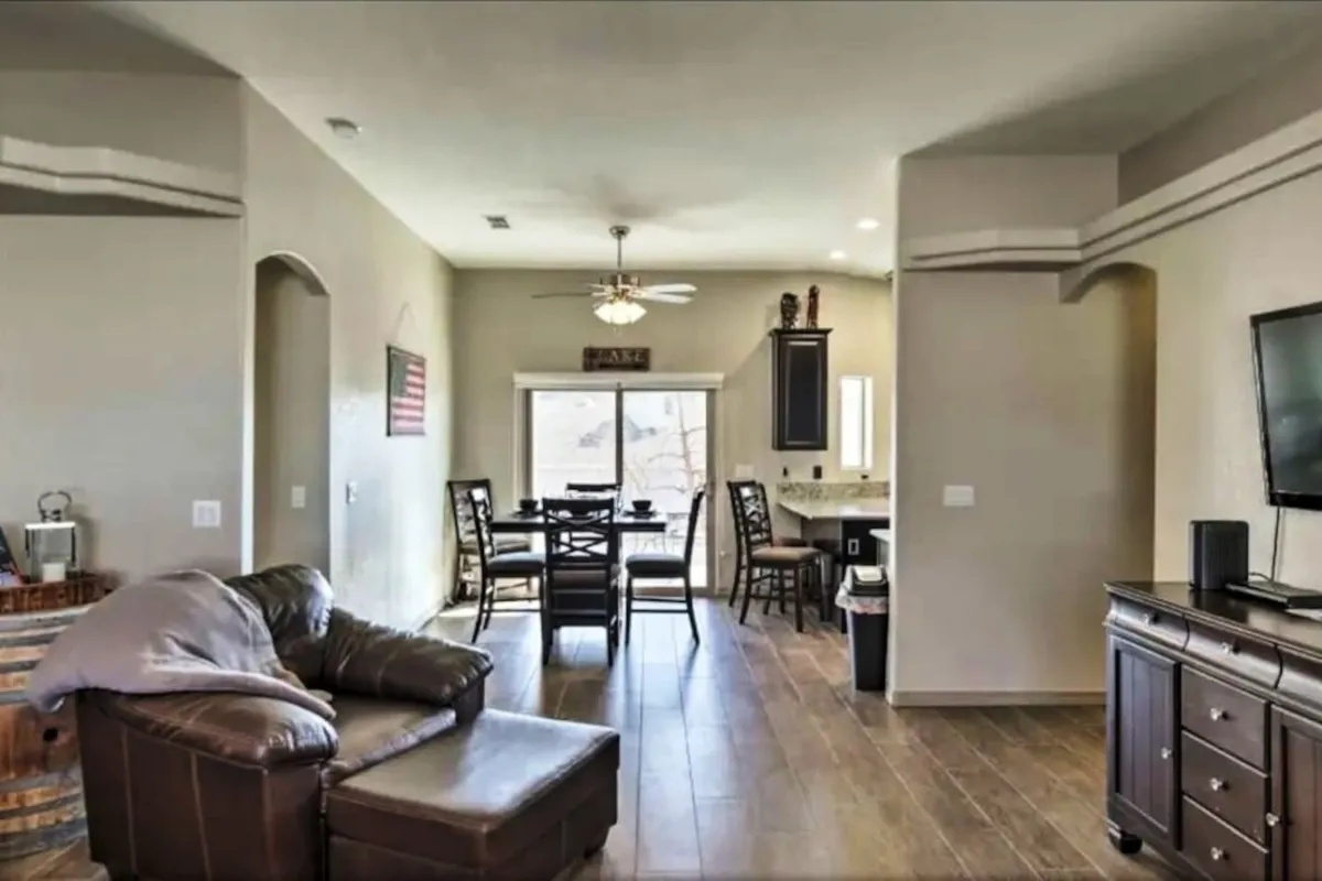 A warm, cozy living space featuring a leather sofa, a prominent dining table, visible kitchen area, and a mounted television. The room is bathed in ambient lighting and set up for both casual dining and relaxed social gatherings.